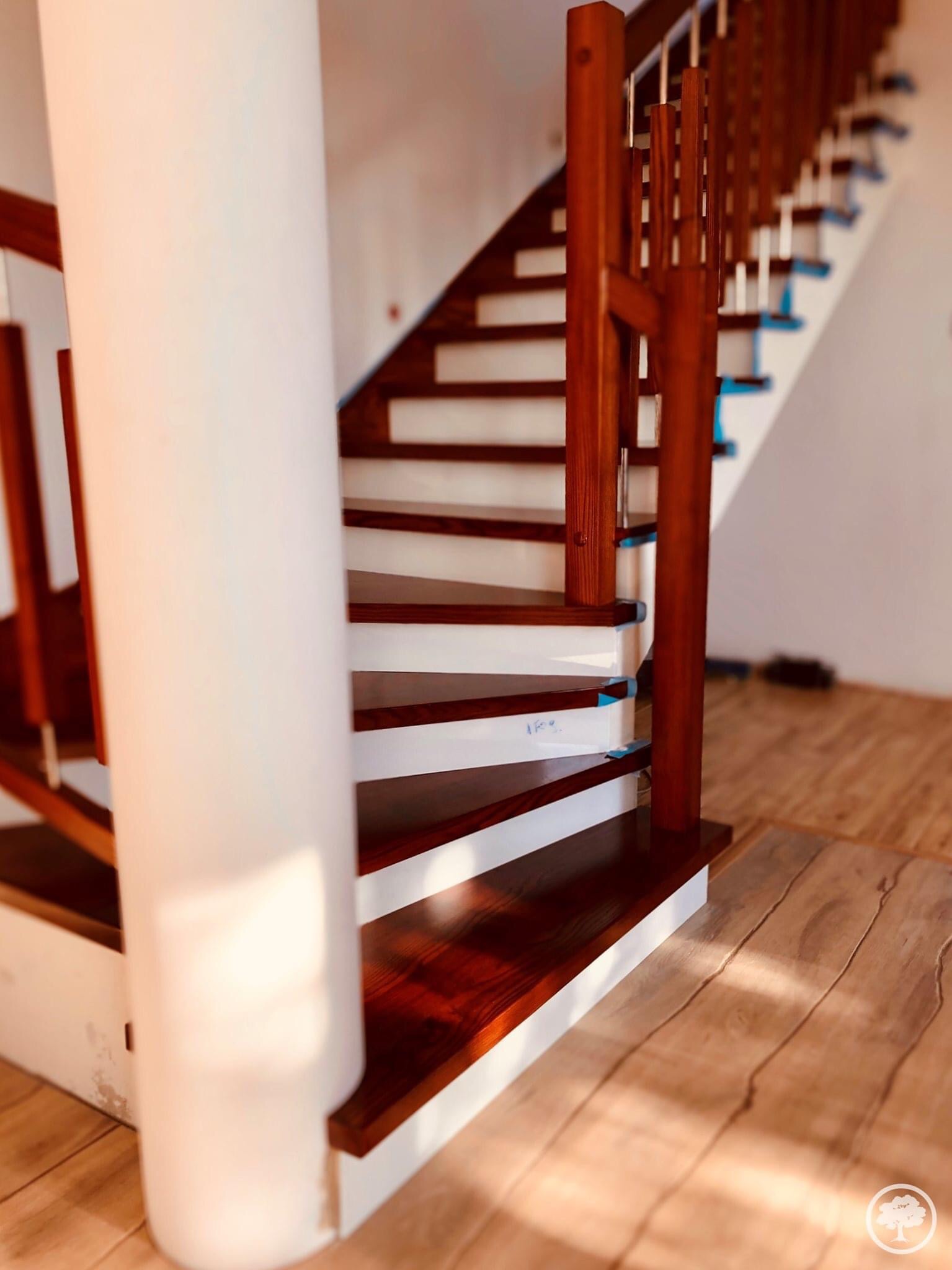 Klatka schodowa z drewnianymi schodami o ciemnym wykończeniu i białych podstopnicach, wsparta na białym, okrągłym filarze, widoczne ślady zabezpieczeń malarskich.