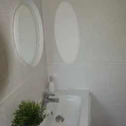 Biała umywalka w łazience z owalnym lustrem, baterią i dozownikiem mydła, uzupełniona o małą doniczkę z zieloną rośliną, na tle białych, wzorzystych płytek.