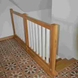 Drewniana barierka schodowa z białymi szczeblami, widoczna na tle wzorzystej, kolorowej podłogi z płytek ceramicznych, przy białej ścianie i schodach.