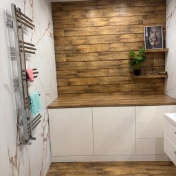 Tomasz Młyński - Nowoczesna łazienka z drewnianą ścianą i blatem, białymi szafkami, marmurowymi płytkami i grzejnikiem drabinkowym z ręcznikami. Uwagę przykuwa obrazek z jeleniem.