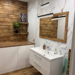 Tomasz Młyński - Nowoczesna łazienka z drewnianymi akcentami: ściana z paneli, blat, rama lustra. Biała szafka pod umywalką, marmurowe płytki i designerskie oświetlenie.