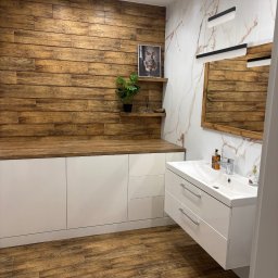 Tomasz Młyński - Nowoczesna łazienka z drewnianą ścianą i podłogą imitującą drewno. Białe szafki pod umywalką i blat z drewnianym dekorem. Marmurowa ściana z lustrem.