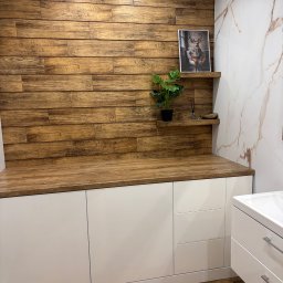 Tomasz Młyński - Białe szafki łazienkowe z drewnianym blatem i panelami ściennymi imitującymi drewno. Na blacie roślina w doniczce i dekoracje. Na ścianie obrazek z jeleniem.