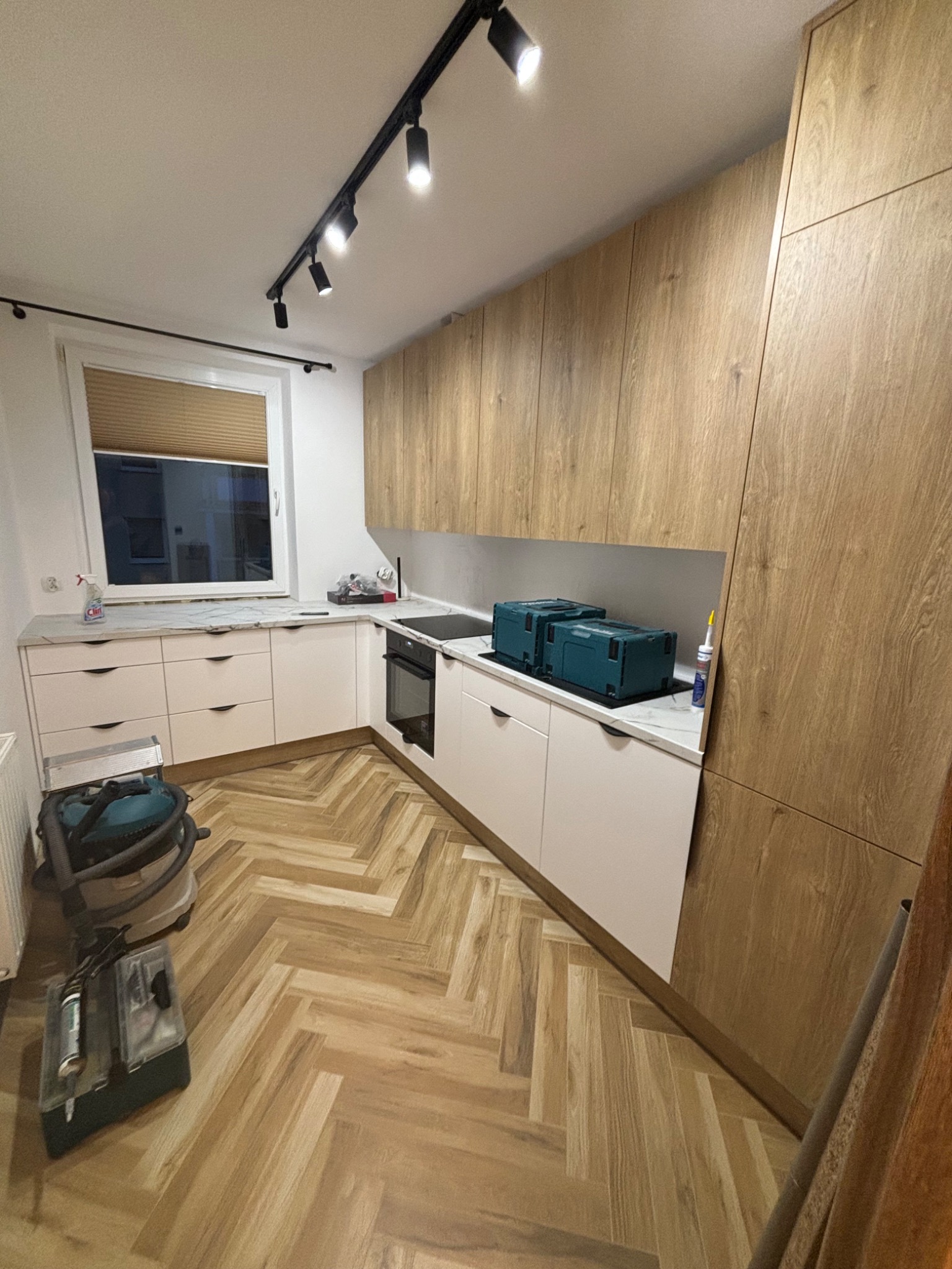 Jasna kuchnia z drewnianymi frontami szafek i białymi blatami. Na podłodze parkiet ułożony w jodełkę. Widoczne narzędzia i akcesoria, co sugeruje trwający montaż.