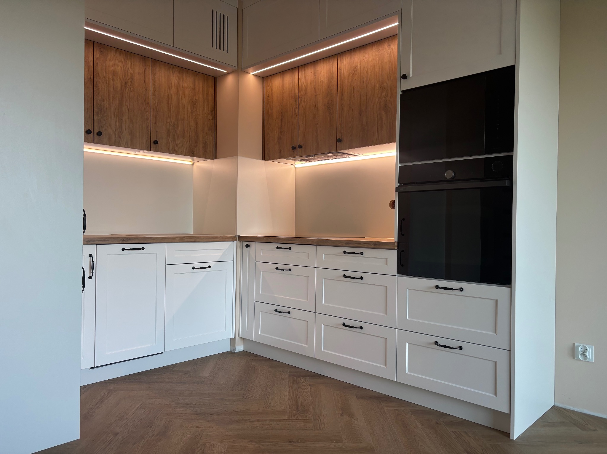 Jasna, narożna kuchnia na wymiar z drewnianym blatem, białymi frontami i oświetleniem LED. Widoczny parkiet w jodełkę i czarny piekarnik w zabudowie.