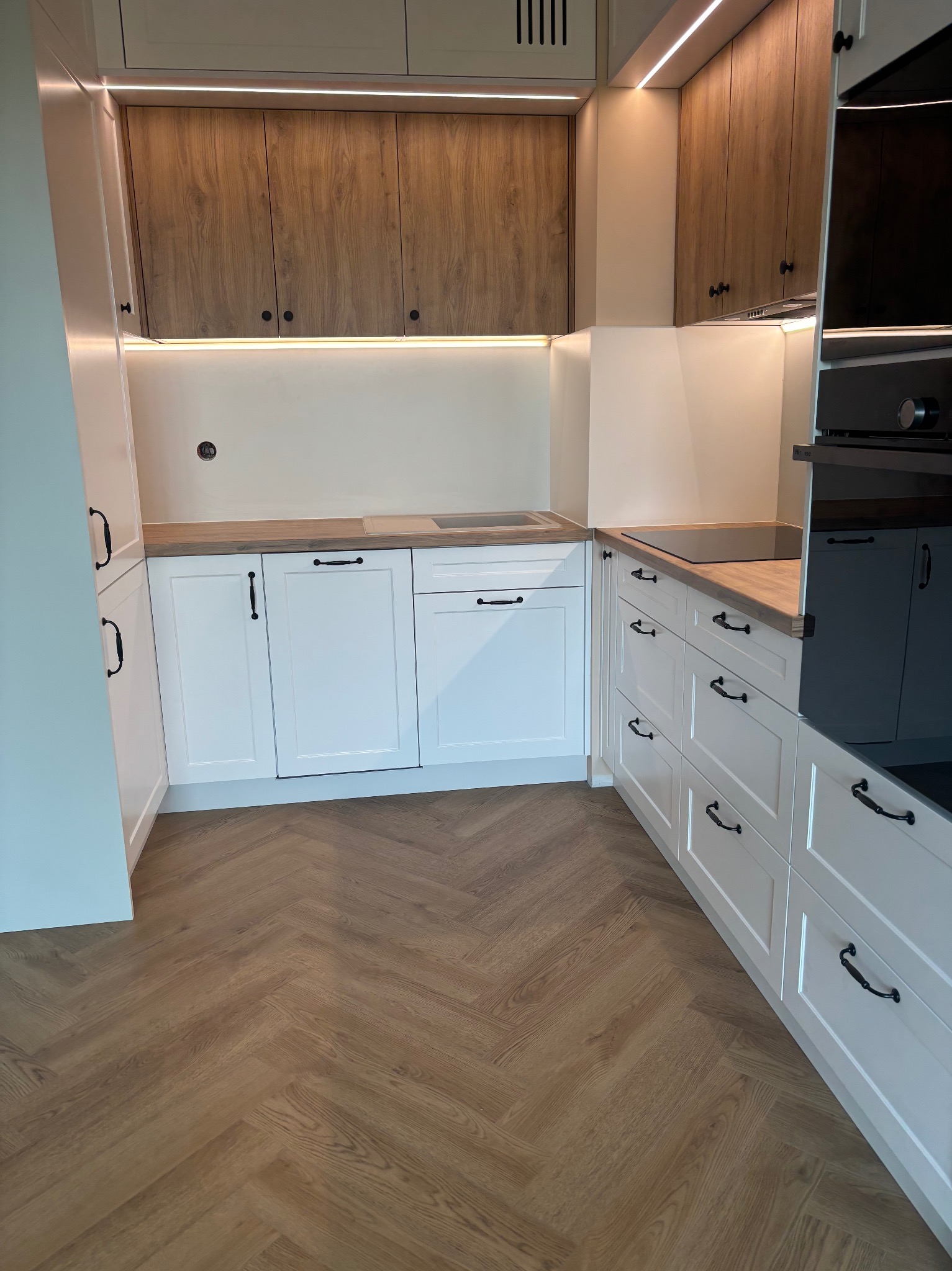 Nowoczesna kuchnia z białymi szafkami i drewnianymi blatami, podświetleniem LED i parkietem w jodełkę. Uwagę zwracają czarne, stylowe uchwyty.