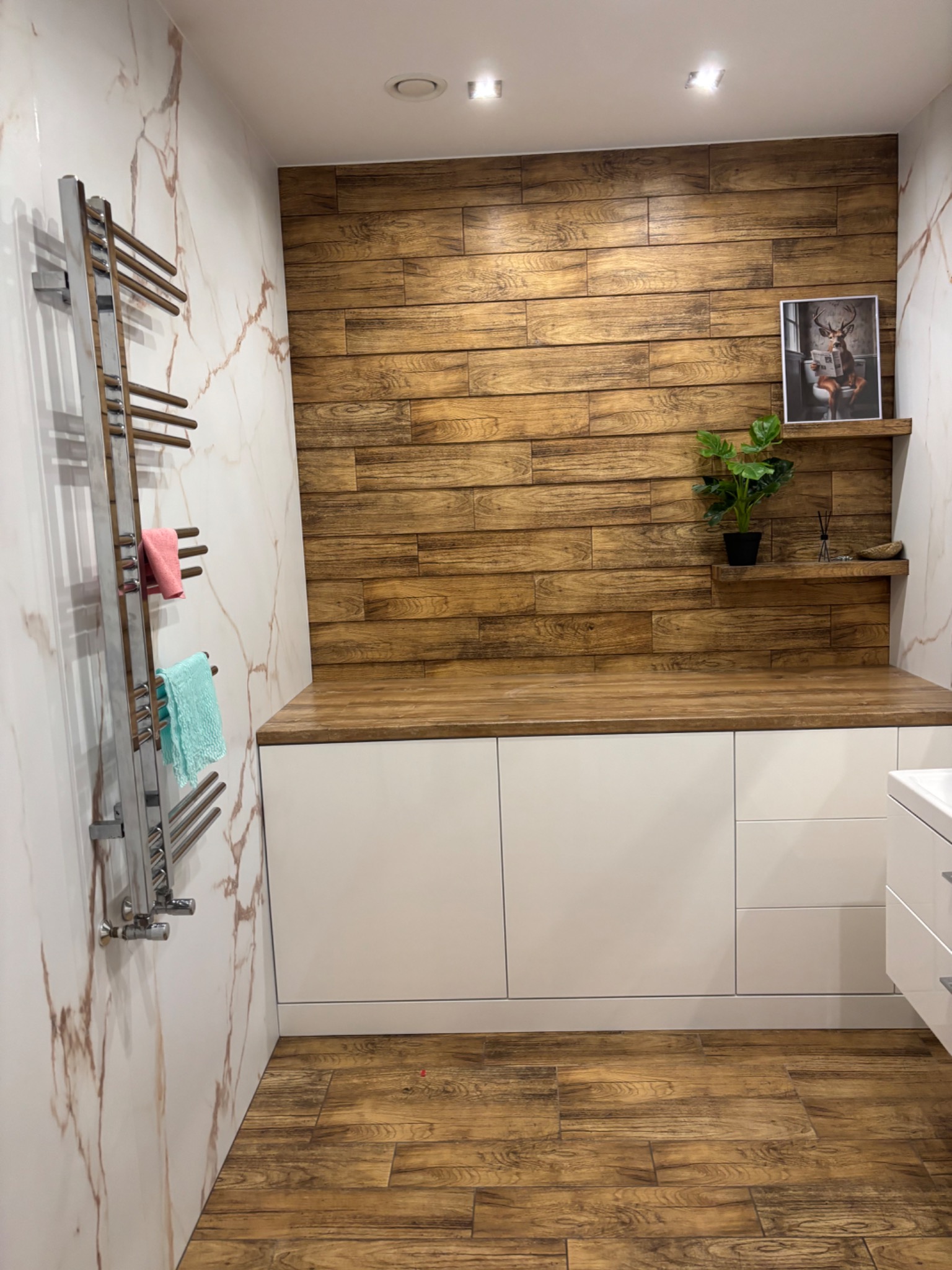 Nowoczesna łazienka z drewnianą ścianą i blatem, białymi szafkami, marmurowymi płytkami i grzejnikiem drabinkowym z ręcznikami. Uwagę przykuwa obrazek z jeleniem.