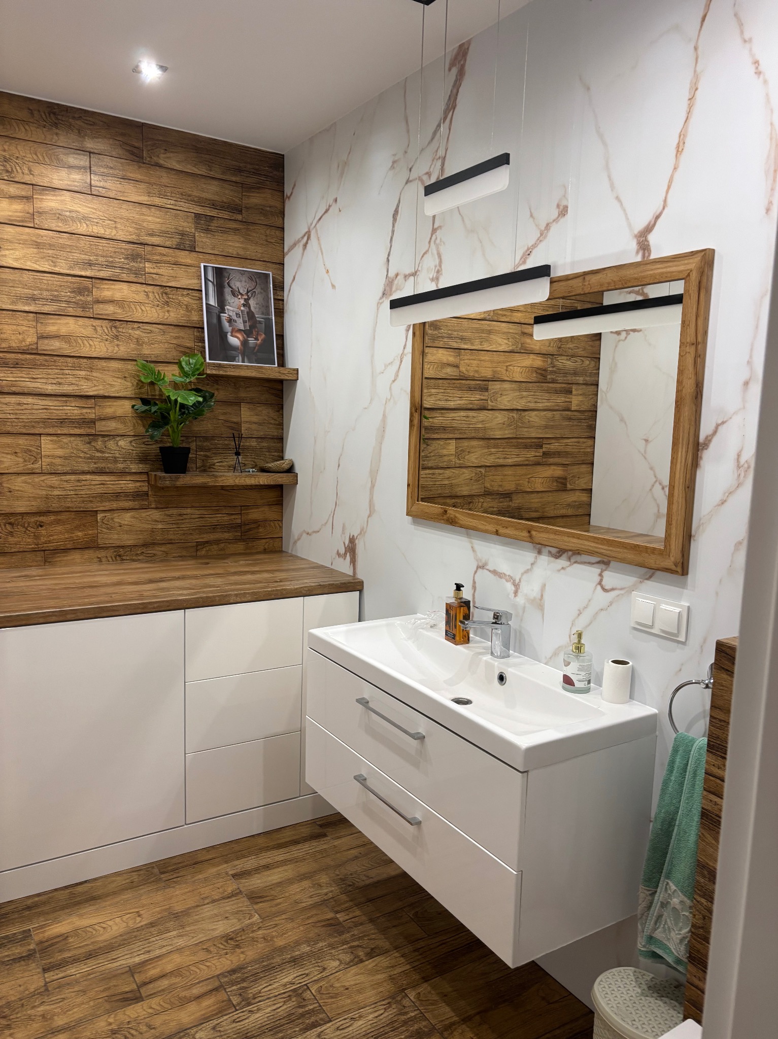 Nowoczesna łazienka z drewnianymi akcentami: ściana z paneli, blat, rama lustra. Biała szafka pod umywalką, marmurowe płytki i designerskie oświetlenie.