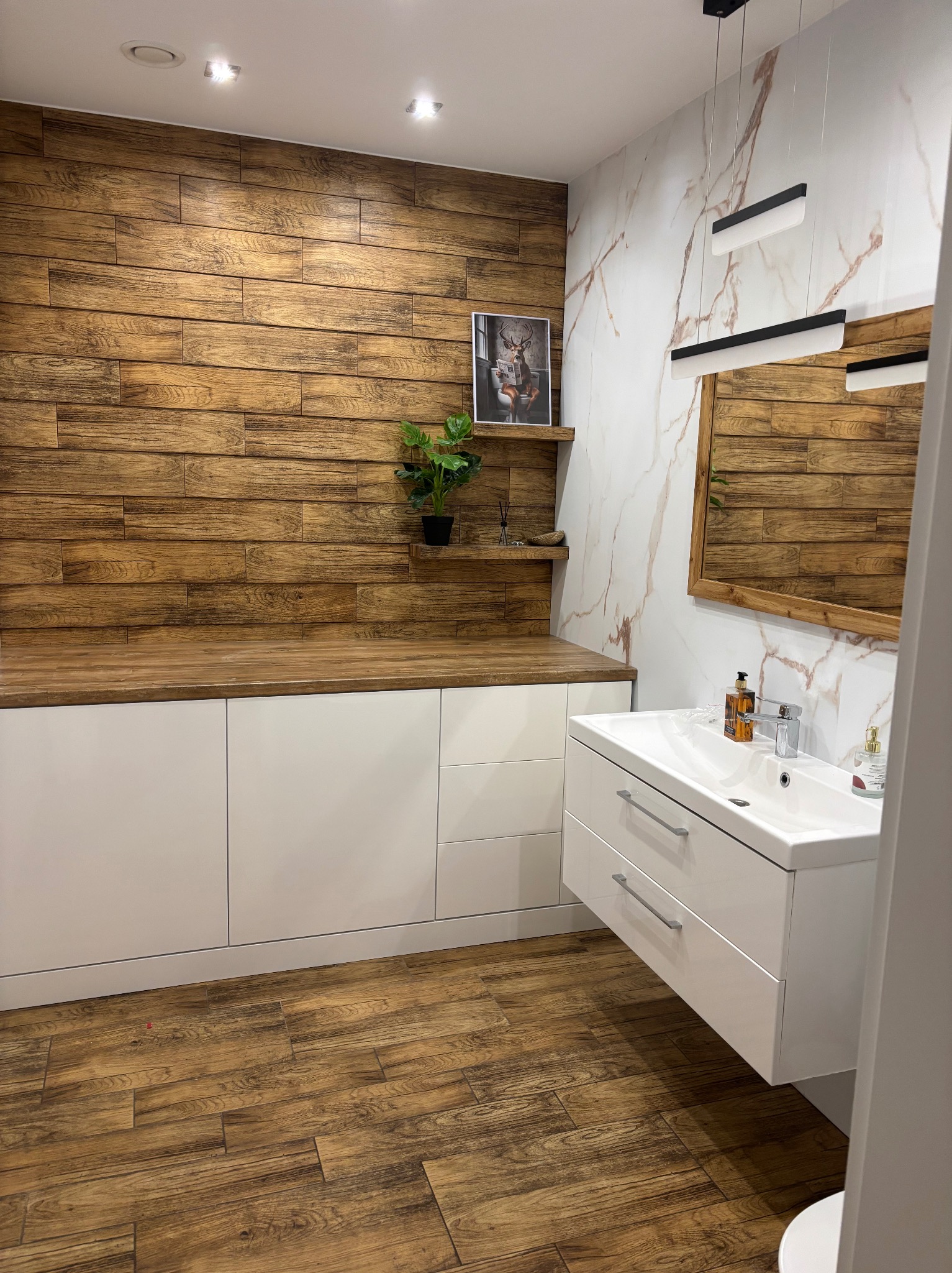Nowoczesna łazienka z drewnianą ścianą i podłogą imitującą drewno. Białe szafki pod umywalką i blat z drewnianym dekorem. Marmurowa ściana z lustrem.