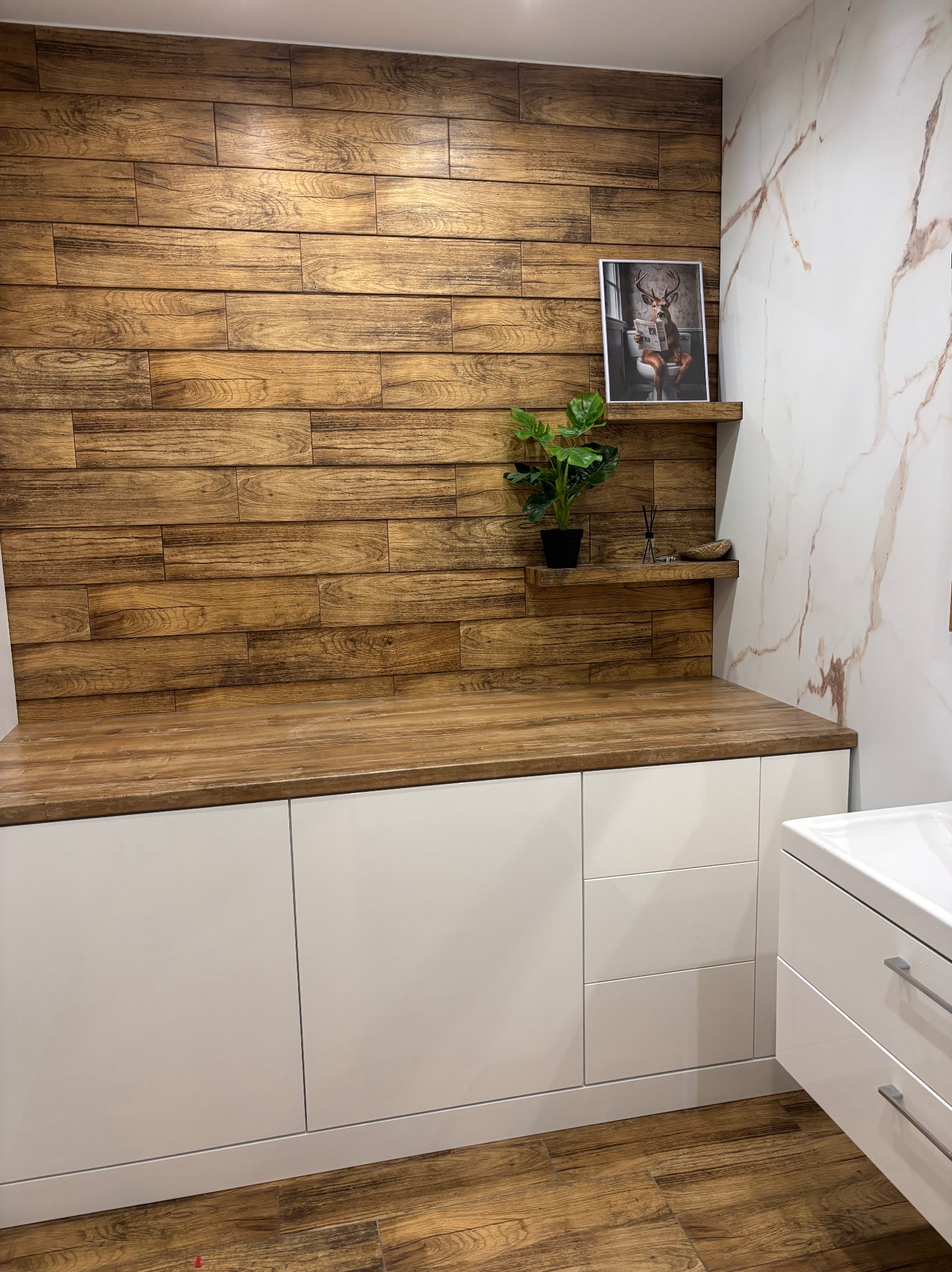 Białe szafki łazienkowe z drewnianym blatem i panelami ściennymi imitującymi drewno. Na blacie roślina w doniczce i dekoracje. Na ścianie obrazek z jeleniem.