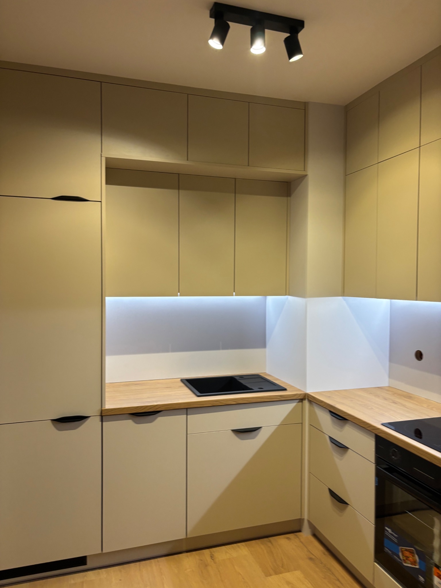Beżowa zabudowa kuchenna z drewnianym blatem i oświetleniem LED w Tczewie. Minimalistyczne uchwyty, nowoczesny design, widoczny fragment płyty indukcyjnej.