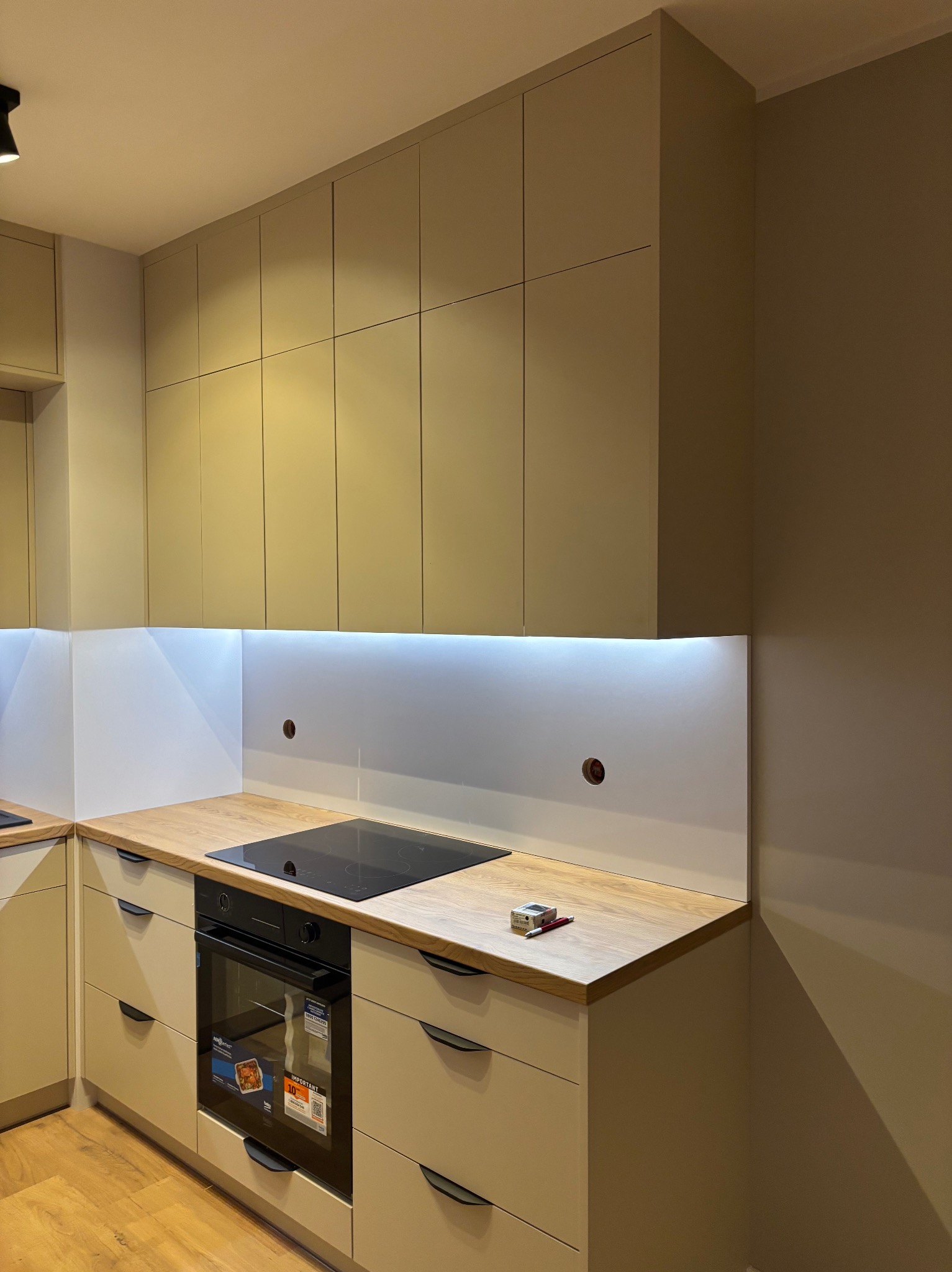 Jasna kuchnia z drewnianym blatem i oświetleniem LED pod szafkami. Widoczna płyta indukcyjna i piekarnik. Na blacie leżą narzędzia. Ujęcie z niskiego kąta.