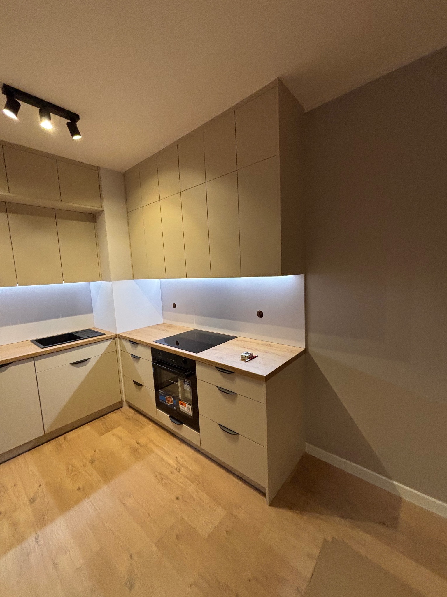 Jasna kuchnia z drewnianym blatem i oświetleniem LED pod szafkami. Na blacie leży miarka i ołówek. Widoczna płyta indukcyjna i piekarnik.