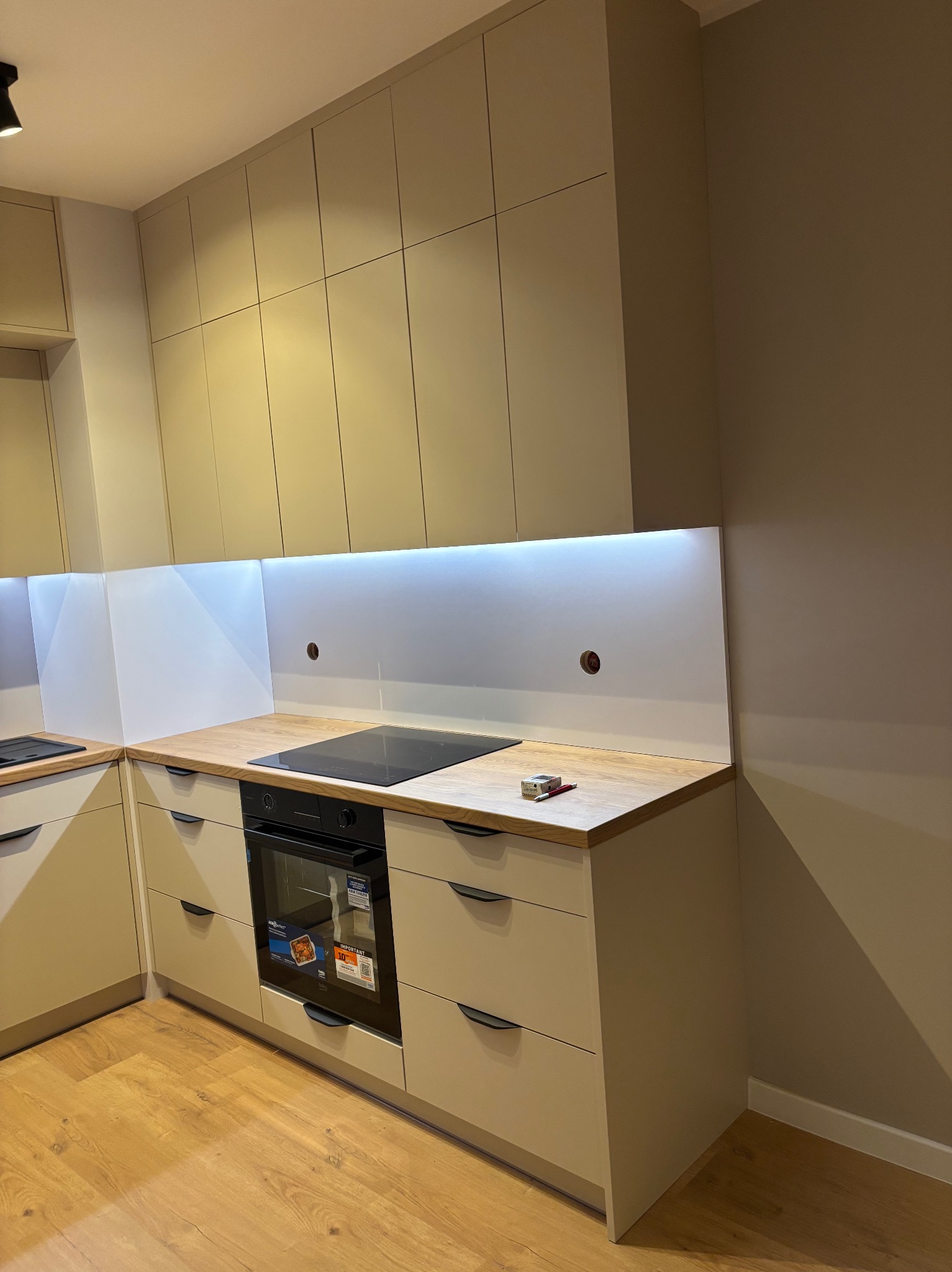 Beżowa kuchnia z drewnianym blatem i oświetleniem LED pod szafkami. Widoczna płyta indukcyjna i piekarnik. Na blacie leżą zapałki i długopis.