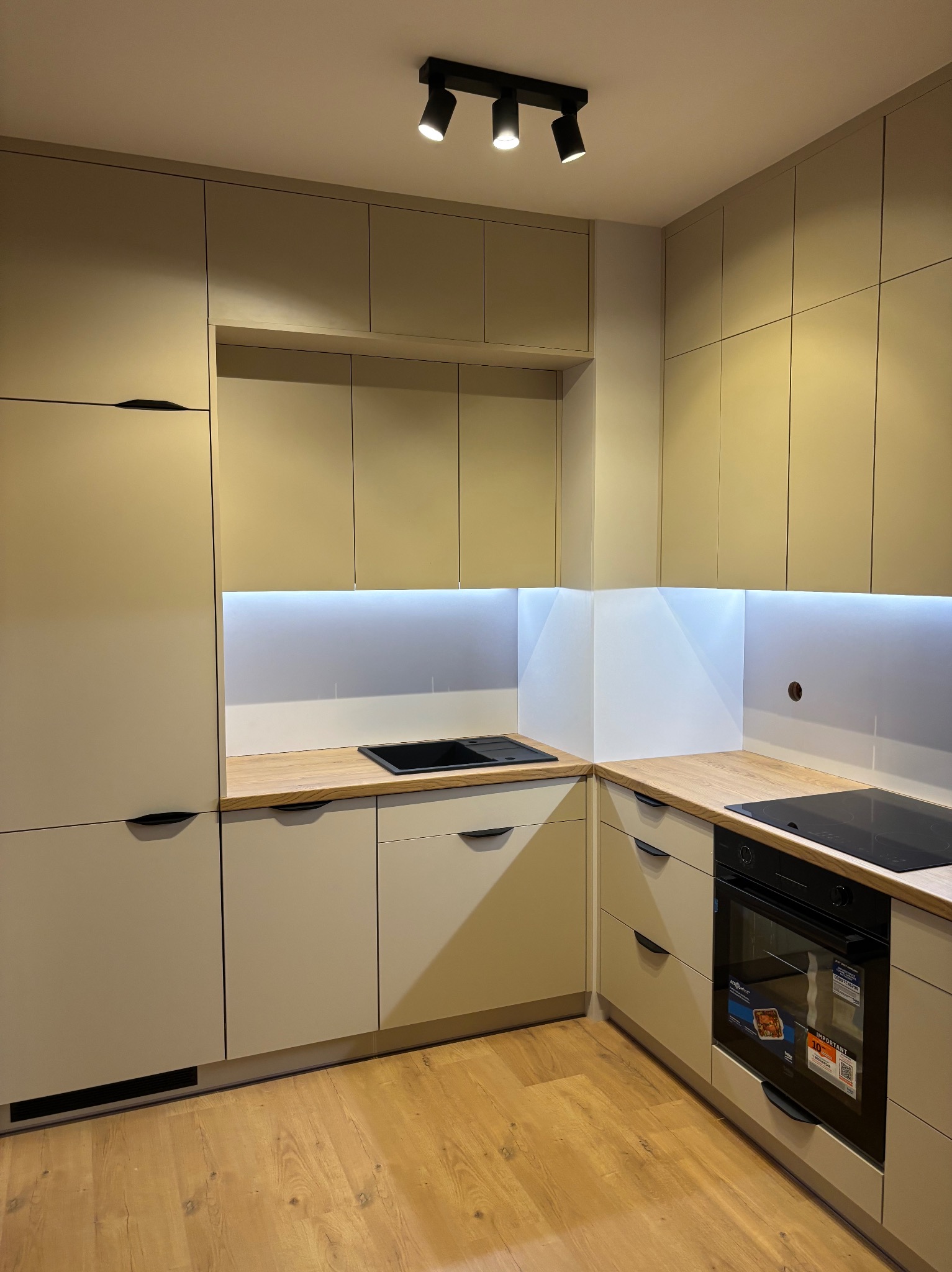 Beżowa kuchnia z drewnianym blatem, oświetleniem LED podszafkowym i czarnymi uchwytami. Widoczny piekarnik i płyta indukcyjna. Minimalistyczny design.