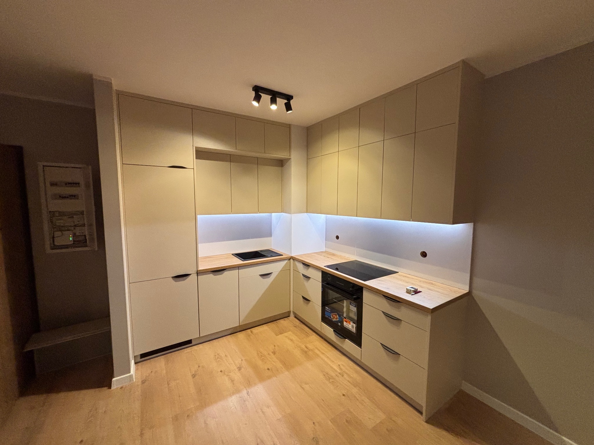 Beżowa kuchnia na wymiar z drewnianym blatem i oświetleniem LED podszafkowym. Widoczne płyta indukcyjna, piekarnik i minimalistyczne uchwyty.