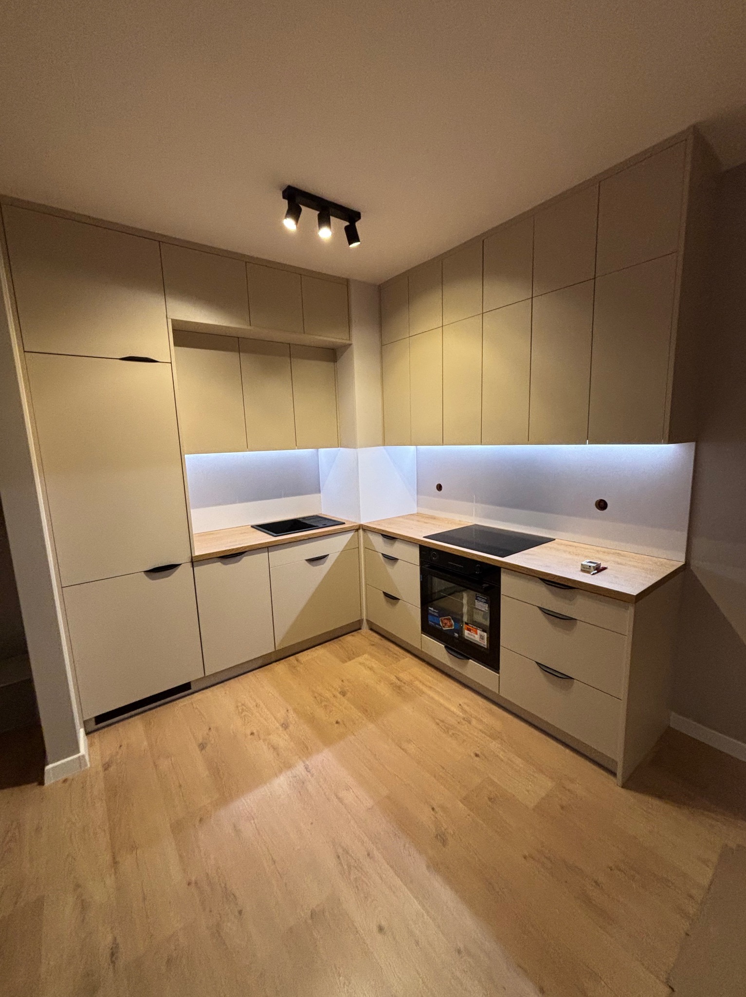 Beżowa kuchnia na wymiar z drewnianym blatem, oświetleniem LED podszafkowym i czarną płytą grzewczą. Ujęcie z niskiego kąta, widoczna podłoga z paneli.