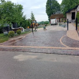 Nowo ułożona, dwukolorowa nawierzchnia z kostki brukowej przed domem, z widocznymi maszynami budowlanymi i przygotowanym terenem, sugerująca trwające prace wykończeniowe.