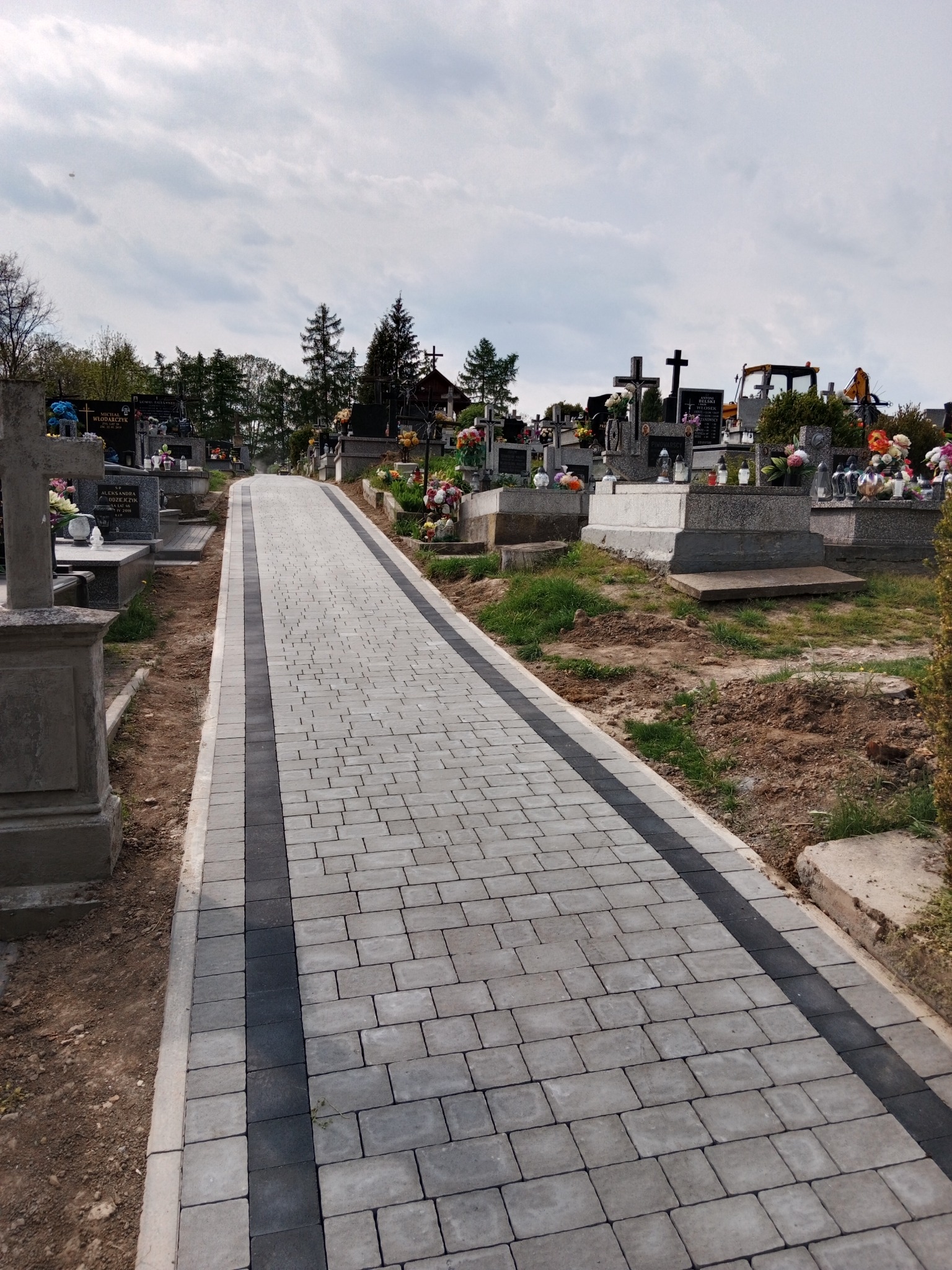 Nowo ułożona ścieżka z szarej kostki brukowej z czarnym pasem pośrodku, prowadząca przez cmentarz między nagrobkami.