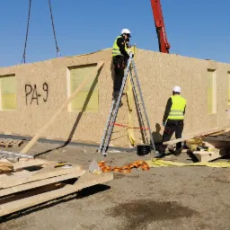Montaż modułu domu z płyty OSB z oznaczeniem PA-9 za pomocą dźwigu, pracownicy w kaskach i kamizelkach odblaskowych, widoczne elementy konstrukcyjne i narzędzia na placu budowy.
