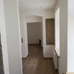 Korytarz w trakcie remontu z ułożoną drewnopodobną podłogą, świeżo pomalowanymi ścianami na biało, grzejnikiem pod oknem i widoczną instalacją elektryczną na suficie.