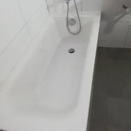 Biała wanna, chromowana bateria z prysznicem, białe płytki ścienne i szare płytki podłogowe w łazience po remoncie.