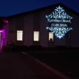 Budynek oświetlony dekoracyjnym projektorem wyświetlającym imiona 'Karolina i Marek' oraz datę 15.09.2018, wejście udekorowane fioletowym oświetleniem i białymi tkaninami.
