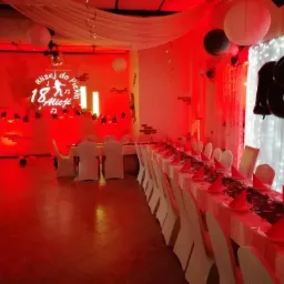 Sala udekorowana na 18-te urodziny Alicji, z projekcją świetlną na ścianie, balonami w kształcie cyfry 18 i długim stołem z czerwonymi serwetkami i białymi pokrowcami na krzesłach, oświetlona...