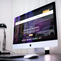 Ekran komputera iMac wyświetlający stronę internetową firmy budowlanej Recabud, z widocznymi elementami interfejsu, lampką biurkową, wazonem z tulipanami i rośliną doniczkową na biurku.