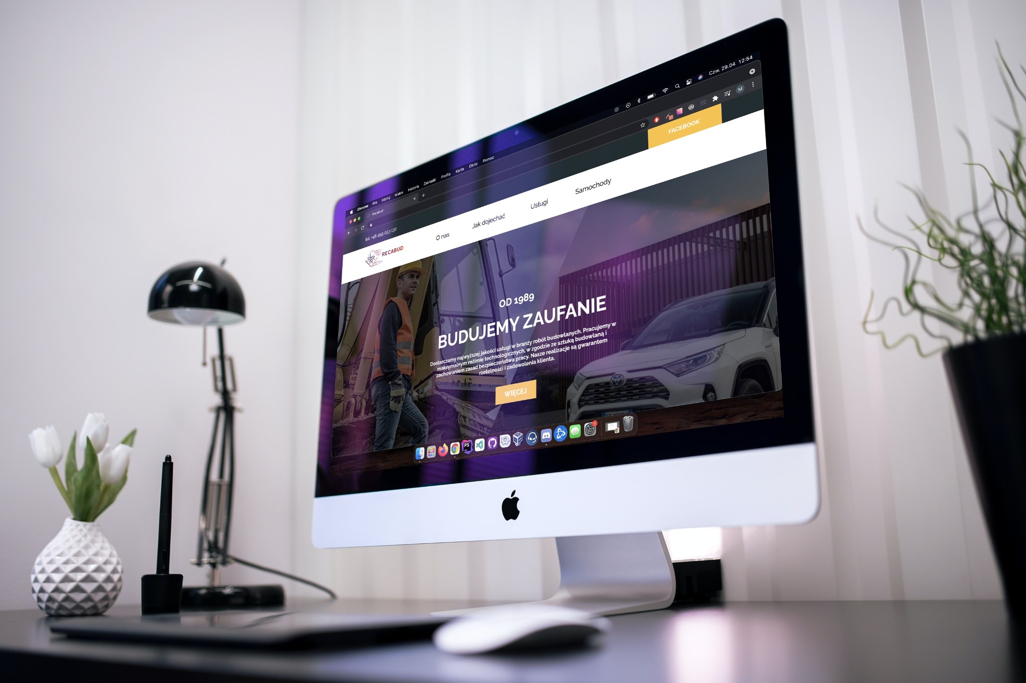 Ekran komputera iMac wyświetlający stronę internetową firmy budowlanej Recabud, z widocznymi elementami interfejsu, lampką biurkową, wazonem z tulipanami i rośliną doniczkową na biurku.
