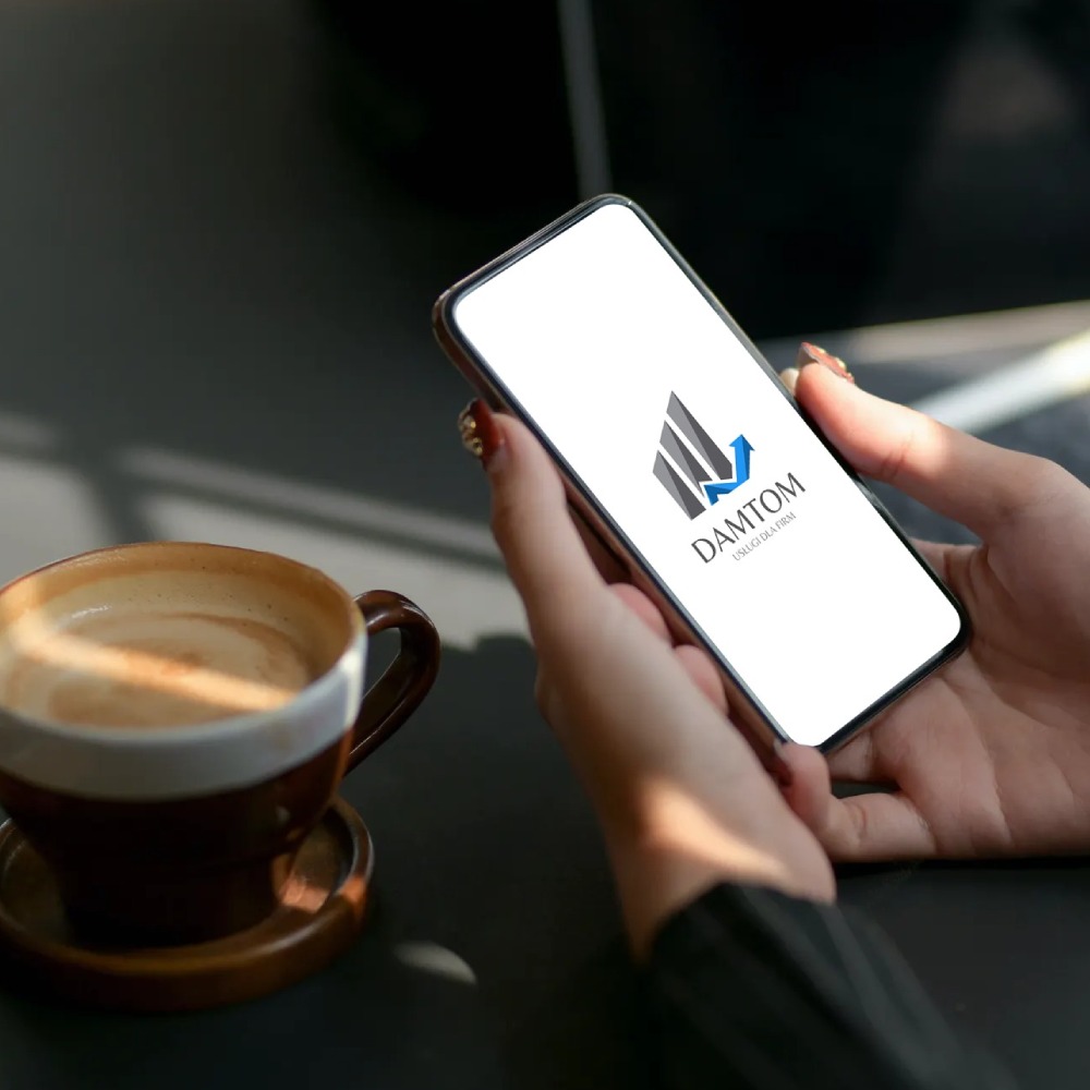Logo firmy DAMTOM wyświetlane na ekranie smartfona trzymanego w dłoniach, obok filiżanka z kawą na ciemnym blacie.