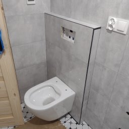 GAC-BUD Usługi Budowlane - Wisząca toaleta w nowoczesnej łazience z szarymi płytkami, widoczne elementy instalacji podtynkowej i fragment podłogi z wzorem geometrycznym.