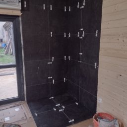 GAC-BUD Usługi Budowlane - Ciemne płytki ułożone na ścianach i podłodze w rogu pomieszczenia z drewnianą boazerią, widoczne krzyżyki dystansowe, wiadro z zaprawą i płytkami na podłodze, okno w tle.