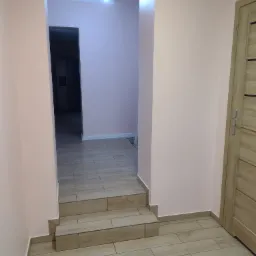 Korytarz z trzema schodkami wyłożonymi płytkami imitującymi drewno, ściany pomalowane na jasny róż, widoczna drewniana ościeżnica drzwi.