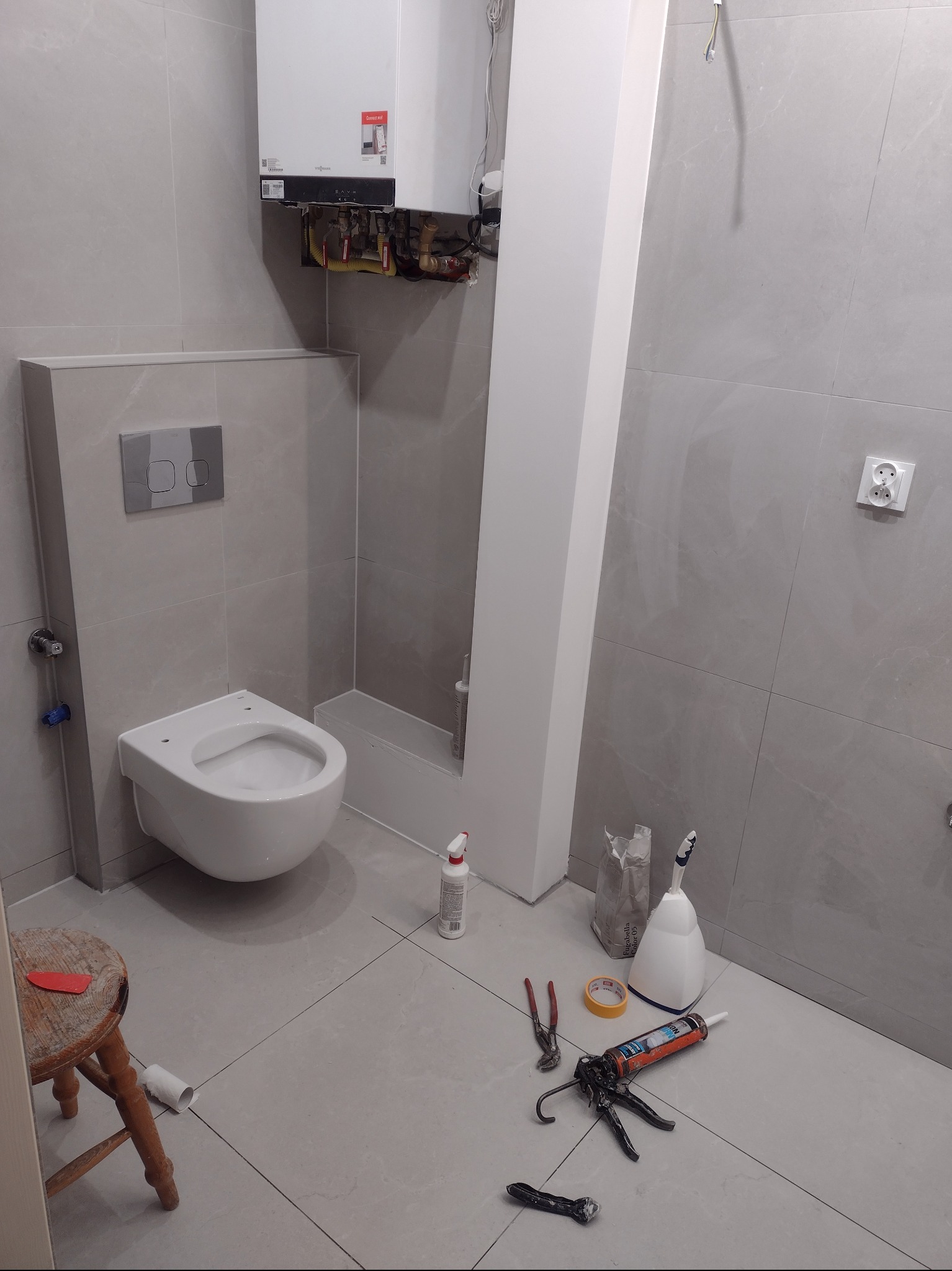 Wisząca toaleta podtynkowa w szarej łazience w trakcie montażu, widoczne narzędzia i materiały budowlane na podłodze, oraz piec gazowy na ścianie.