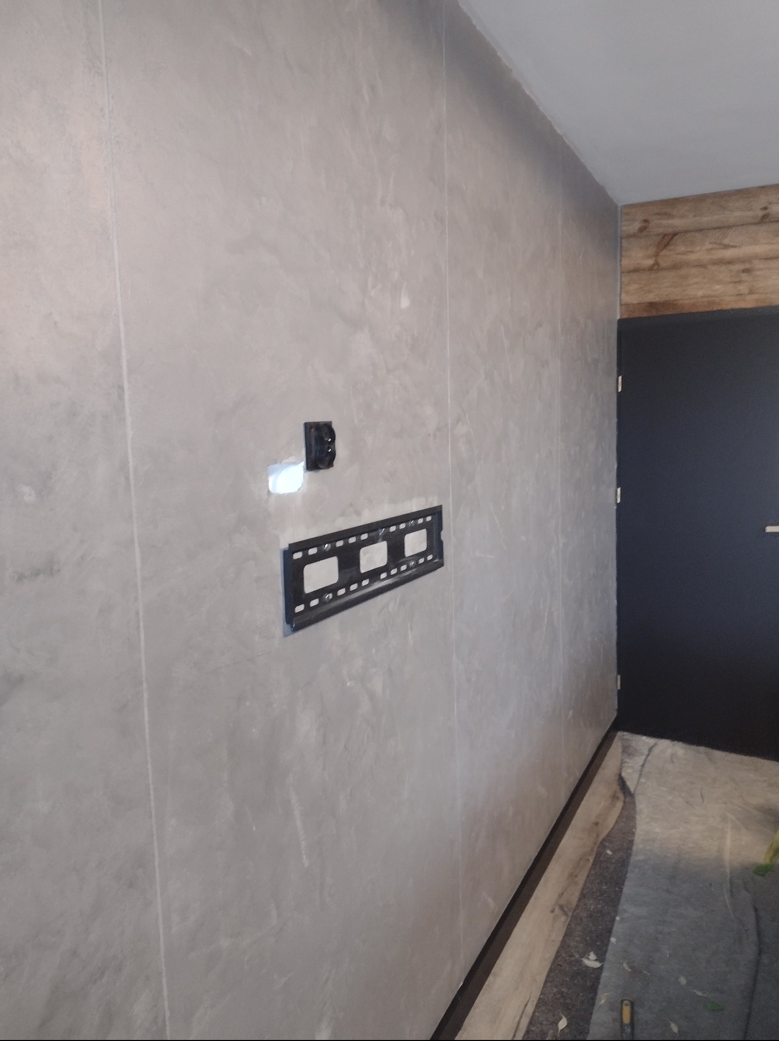 Ściana wykończona szarym tynkiem dekoracyjnym z zamontowanym uchwytem do telewizora i gniazdkiem elektrycznym, widoczny fragment drewnianej ściany i czarne drzwi.