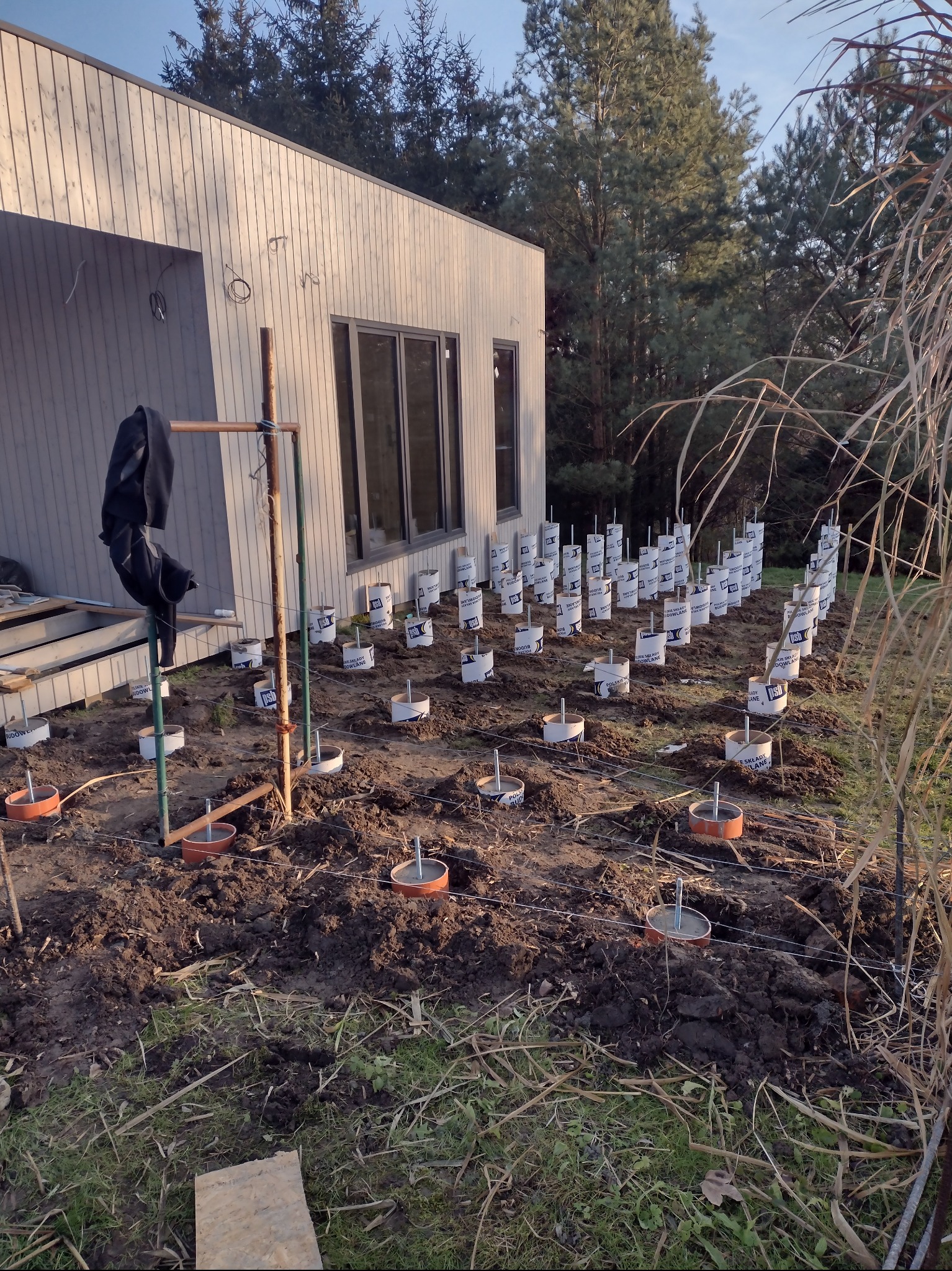 Przygotowanie podłoża pod budowę domu z drewnianą elewacją: rozmieszczone i wypoziomowane słupki fundamentowe z metalowymi trzpieniami, widoczne narzędzia i liny pomiarowe.