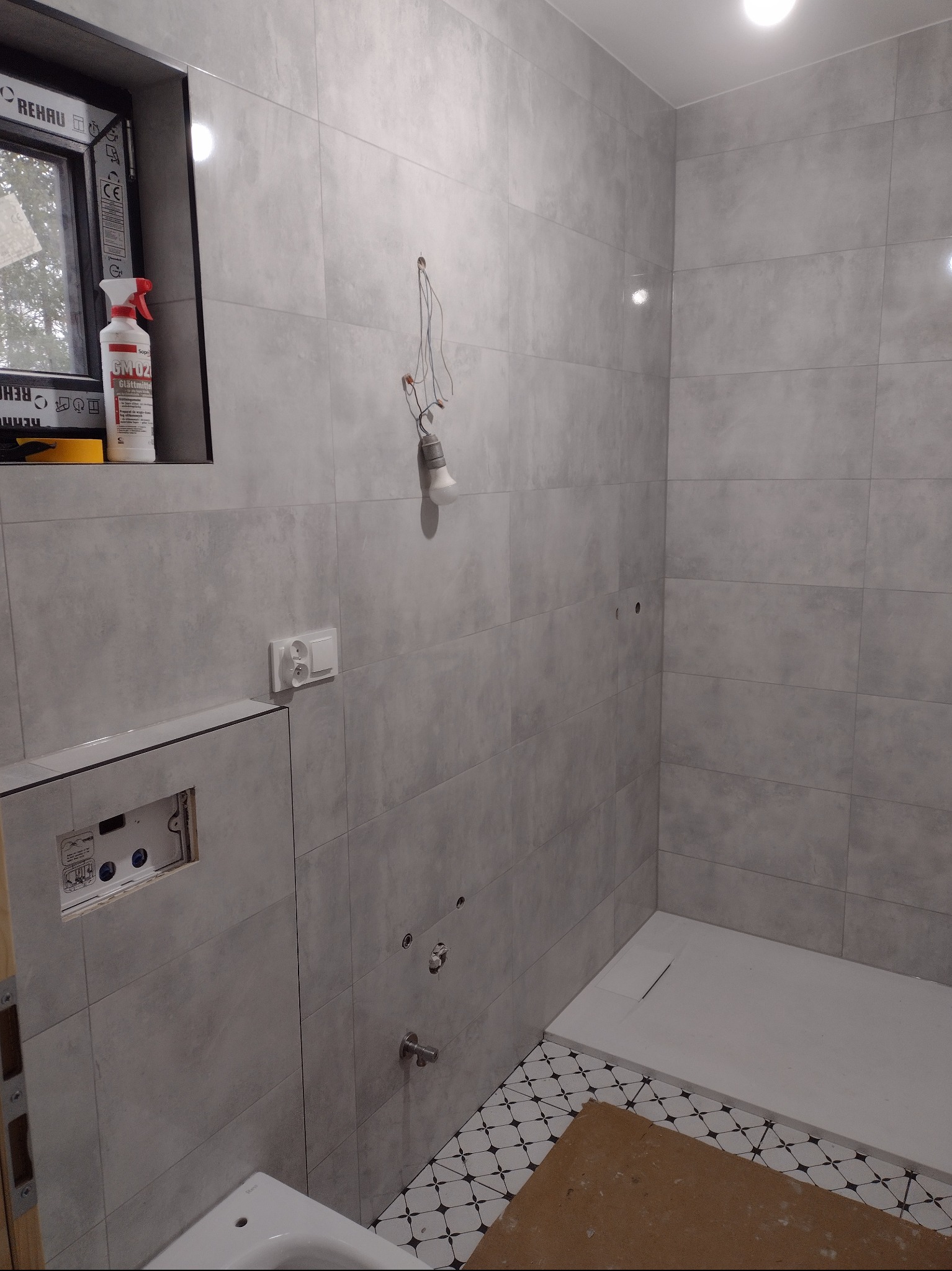 Wnętrze łazienki w trakcie remontu w Wąbrzeźnie: szare, matowe płytki na ścianach, fragment podłogi z czarno-białą mozaiką, widoczna instalacja elektryczna i hydrauliczna przed montażem armatury...