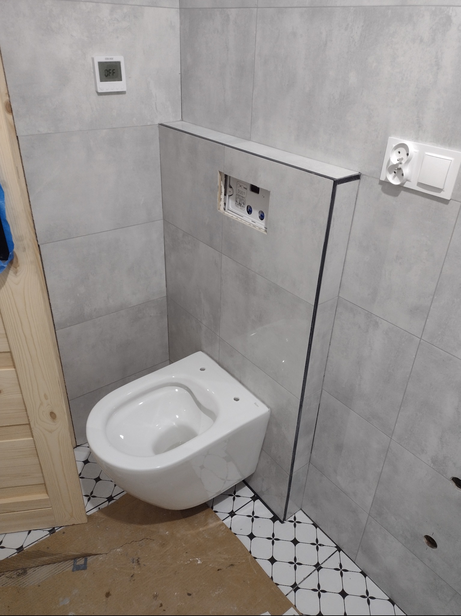 Wisząca toaleta w nowoczesnej łazience z szarymi płytkami, widoczne elementy instalacji podtynkowej i fragment podłogi z wzorem geometrycznym.