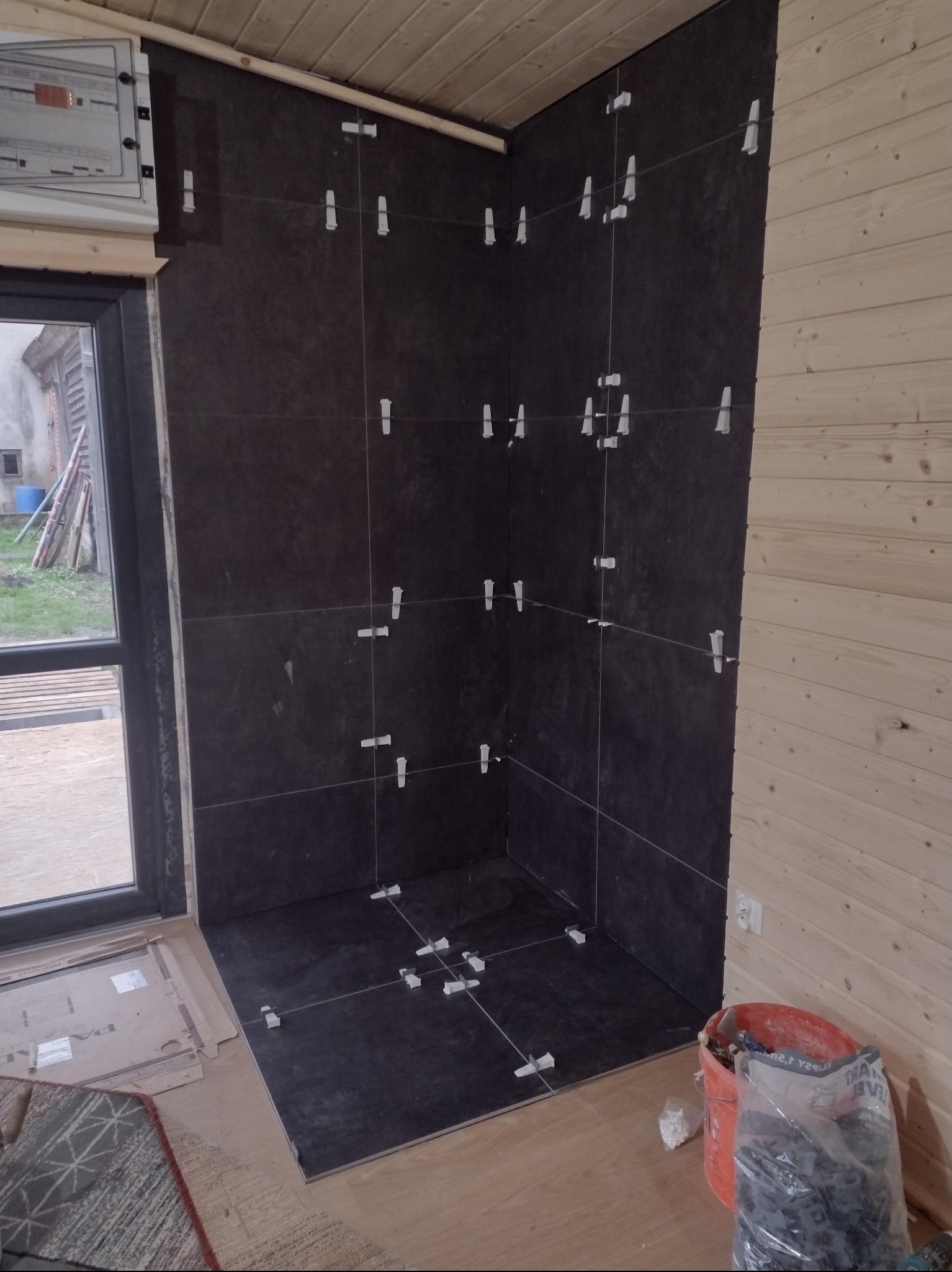 Ciemne płytki ułożone na ścianach i podłodze w rogu pomieszczenia z drewnianą boazerią, widoczne krzyżyki dystansowe, wiadro z zaprawą i płytkami na podłodze, okno w tle.