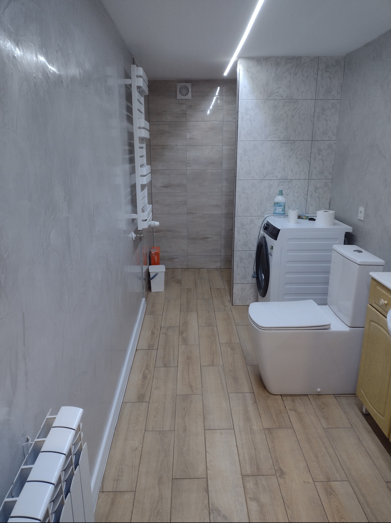 Umywalka, toaleta, pralka i grzejnik w wykończonej łazience z beżowymi panelami podłogowymi i płytkami ściennymi, oświetlenie LED na suficie.