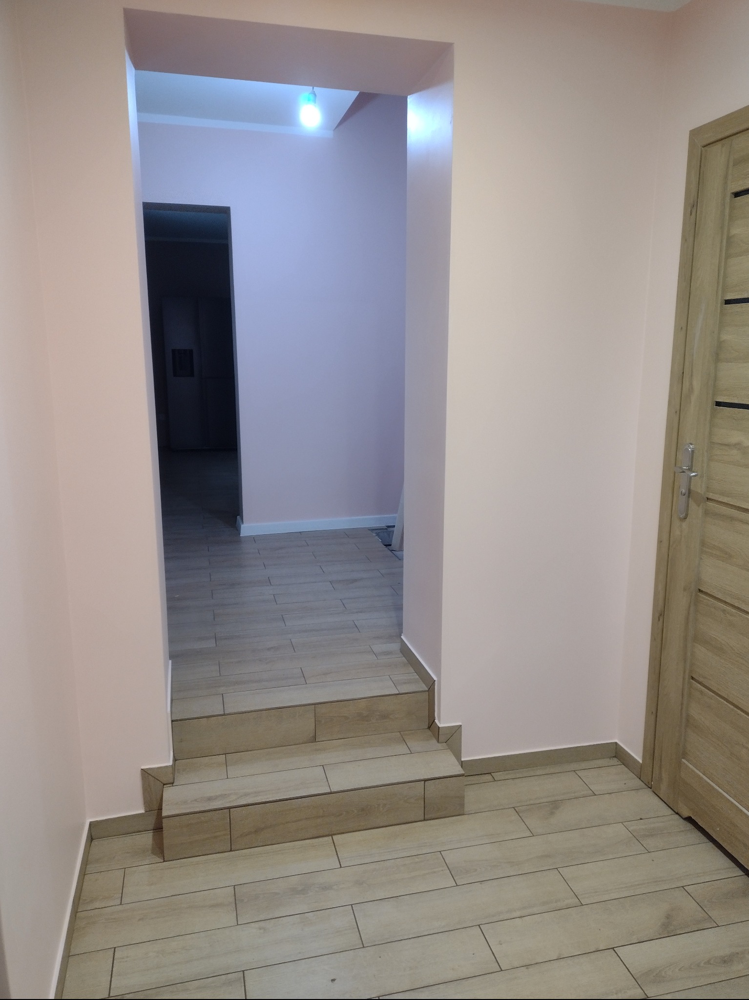 Korytarz z trzema schodkami wyłożonymi płytkami imitującymi drewno, ściany pomalowane na jasny róż, widoczna drewniana ościeżnica drzwi.