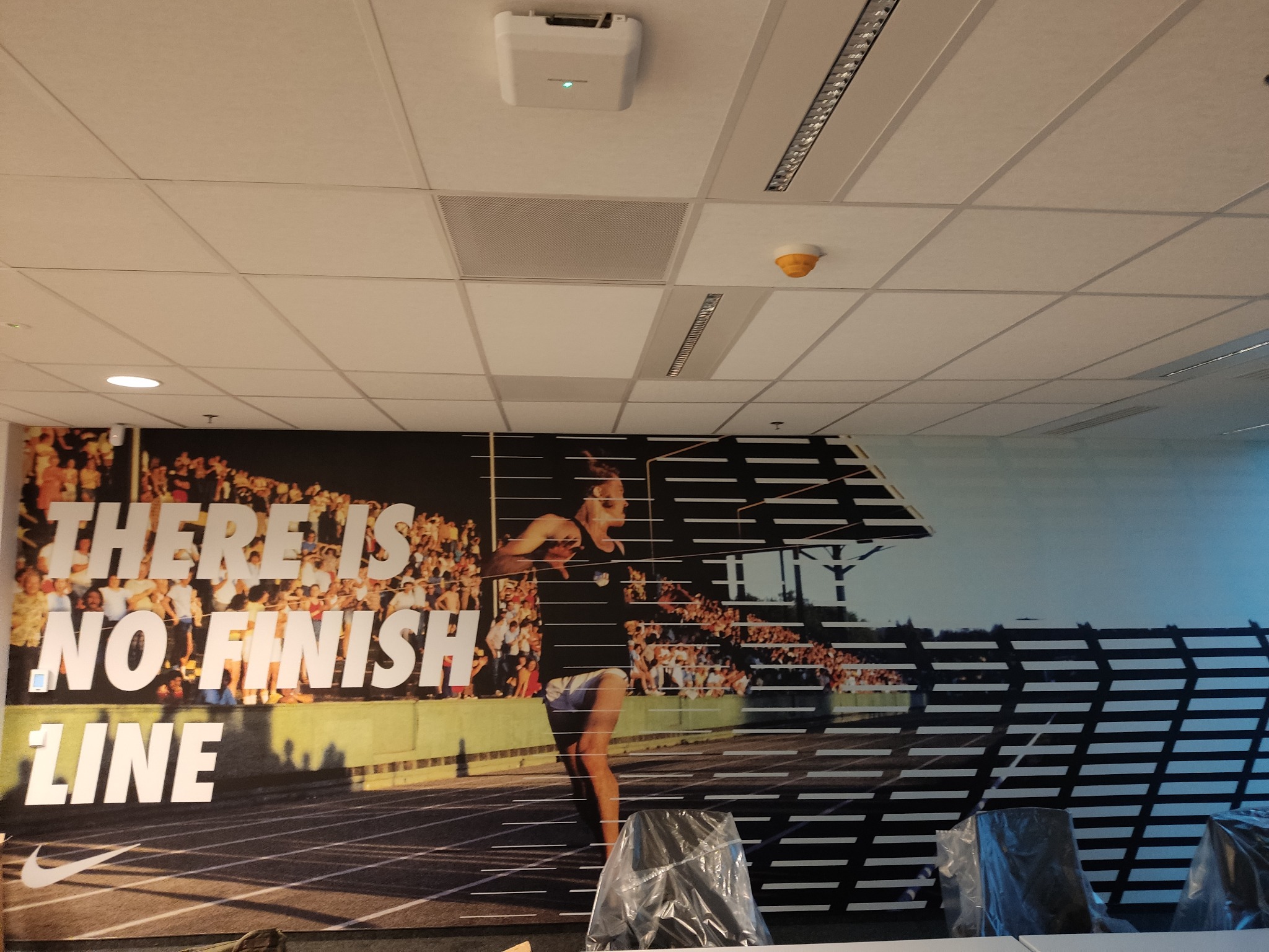 Biuro z fototapetą przedstawiającą biegaczkę i hasło 'There is no finish line' oraz logo Nike, widok na sufit z oświetleniem i klimatyzacją.