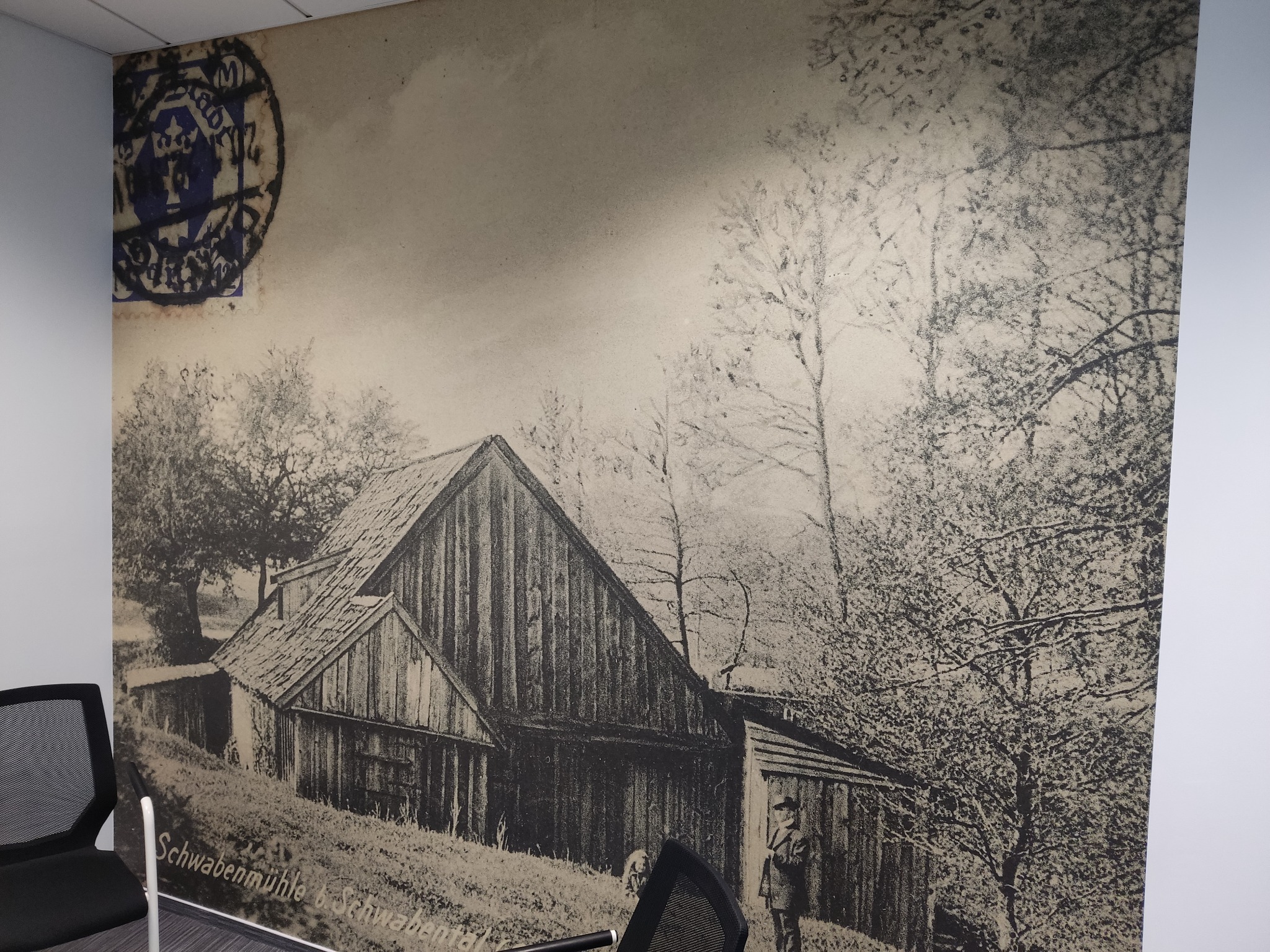 Duża fototapeta przedstawiająca czarno-białą pocztówkę ze stemplem, ukazującą starą drewnianą chatę i drzewa, częściowo zasłonięta przez nowoczesne krzesło biurowe.