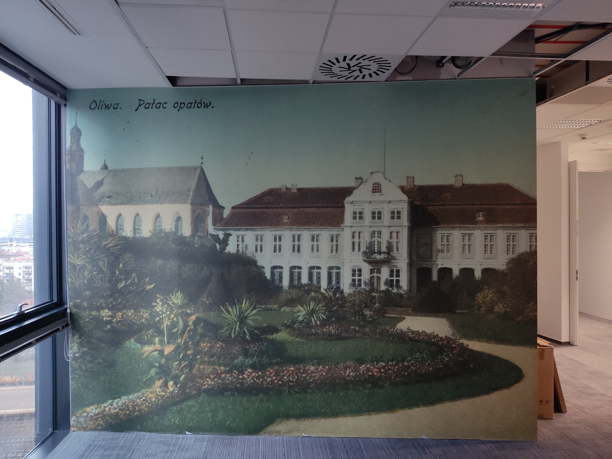 Duża fototapeta przedstawiająca Pałac Opatów w Oliwie, umieszczona w nowoczesnym biurze z widokiem na miasto przez okno.