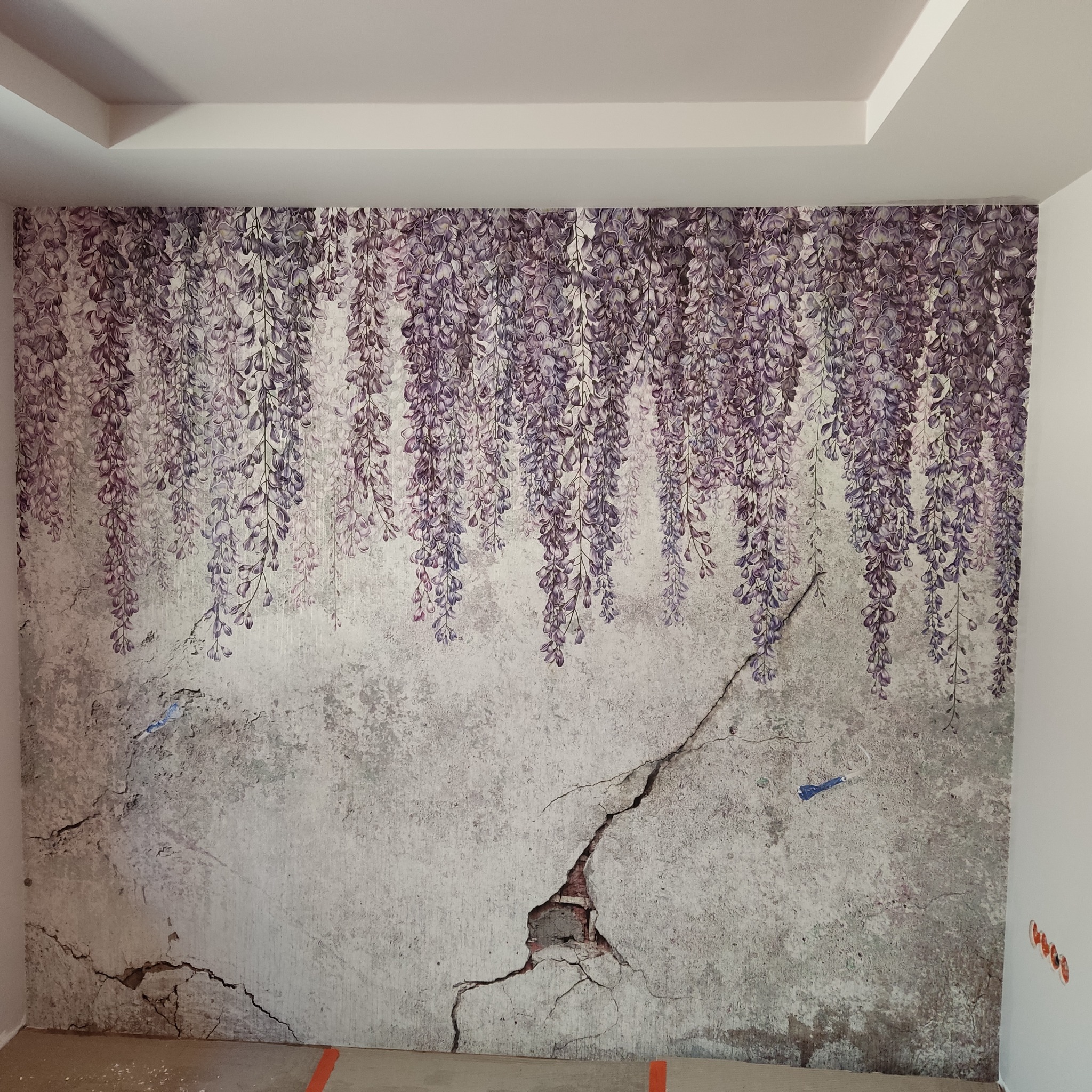 Ściana po nałożeniu tapety z motywem kwitnącej glicynii, imitującej spękaną, starą strukturę muru z widocznymi uszkodzeniami i pęknięciami. Fragment ściany z widocznymi cegłami w miejscu pęknięcia.