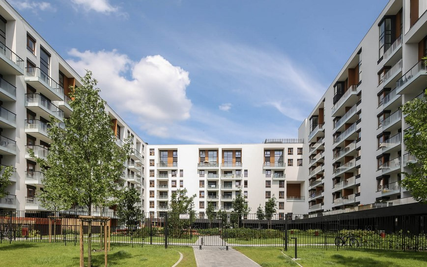 Nowoczesny kompleks apartamentowy z białymi elewacjami, szklanymi balkonami i zagospodarowanym zielonym dziedzińcem wewnętrznym w słoneczny dzień.