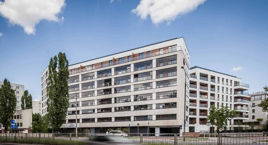 Elewacja nowoczesnego apartamentowca z balkonami, jasna kolorystyka, duże okna, widok z poziomu ulicy, błękitne niebo z chmurami w tle.