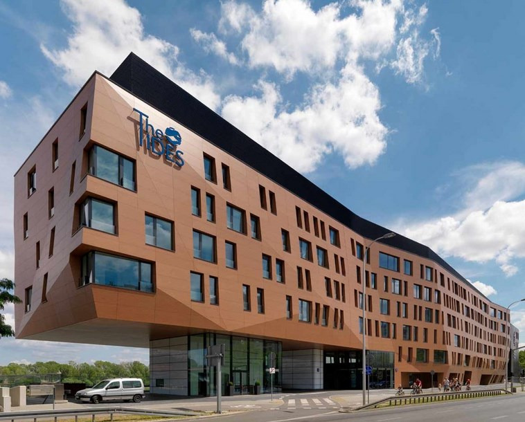 Nowoczesny budynek biurowy z elewacją w odcieniach brązu i dużymi oknami, podświetlony w słoneczny dzień z błękitnym niebem i białymi chmurami w tle.