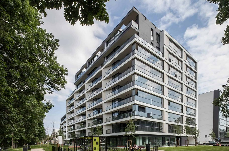 Nowoczesny budynek apartamentowy z balkonami, jasna elewacja z dużymi oknami, widok z poziomu parku z drzewami i elementami małej architektury.